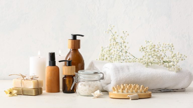 Set de productos naturales para el cuidado personal con frascos, sales de baño, jabones artesanales, cepillo de masaje, toalla blanca y flores decorativas sobre fondo neutro
